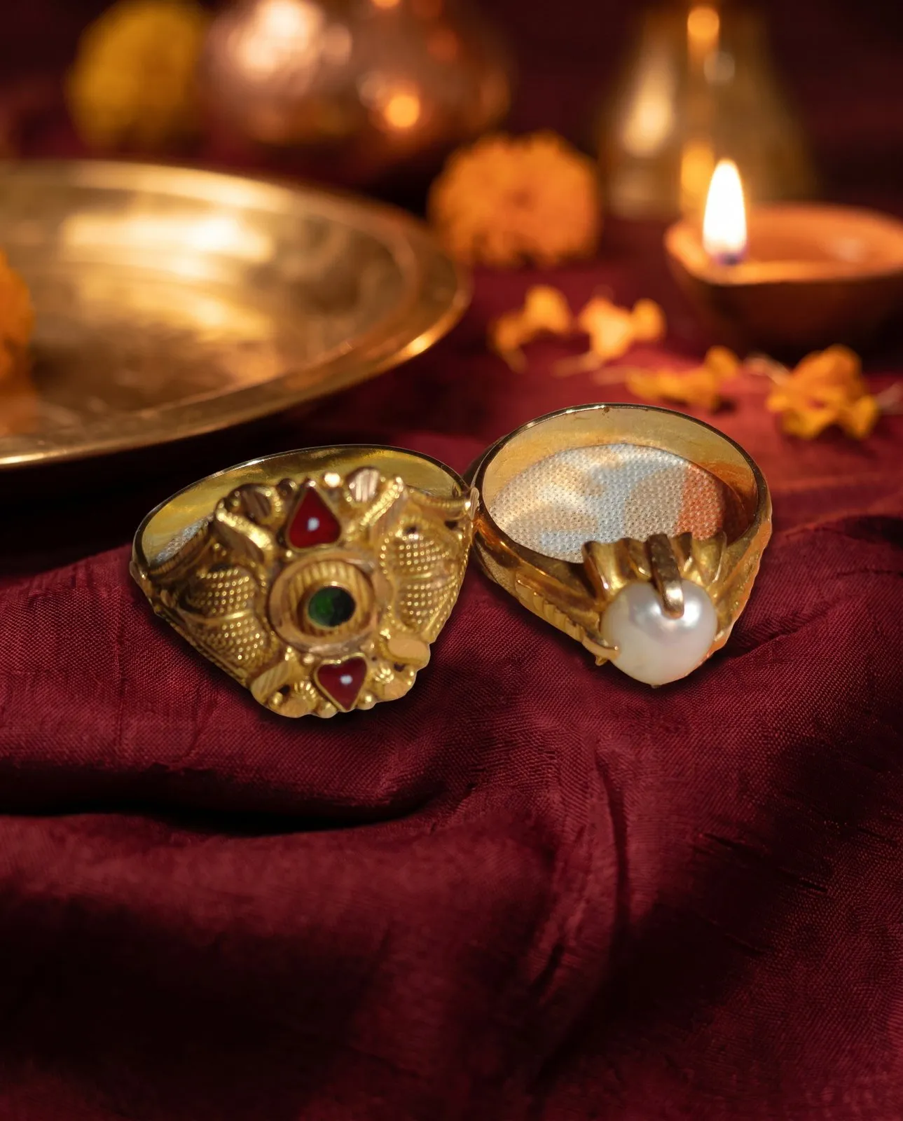 The same two gold rings on maroon silk with a brass diya plate and marigolds behind them